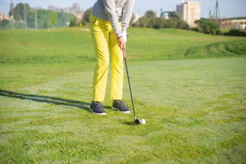 A golfer wearing yellow pants practices putting on a sunny day on a lush green course