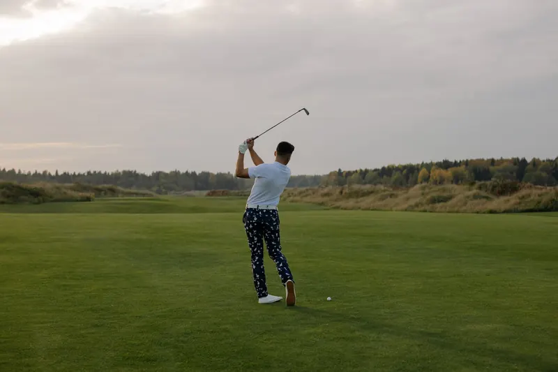 A male golfer in patterned pants swinging a club on a lush green field during sunset